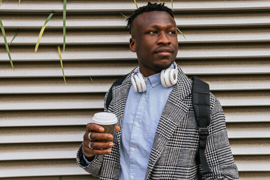 Young contemplative black male office employee with headset and takeaway coffee looking away near ribbed wall in city