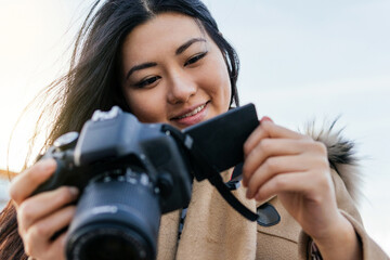 Ethnic young happy Asian female photographer shooting photo on professional photo camera on city street