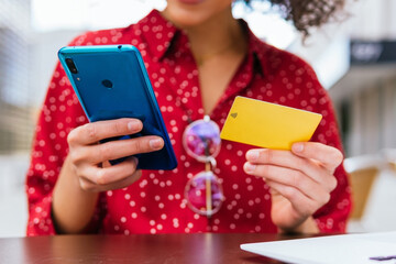 Crop anonymous female in stylish outfit performing online transaction with credit card and mobile phone while sitting at table on cafe terrace