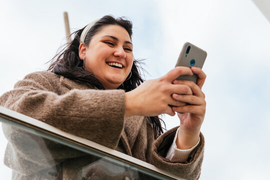 Low angle of young cheerful plump female in coat chatting on cellphone while leaning on fence in city
