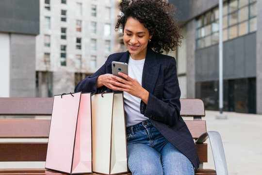 Positive black female sitting on bench with paper bags after shopping and browsing mobile phone in city