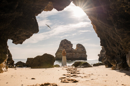 Back view of unrecognizable female in casual clothes and hat standing on sand in entrance of cave near sea looking at a bird in Algarve, Portugal