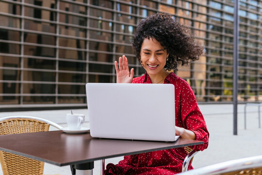 Happy young ethnic female waving hand while communicating with friend during video conference via laptop at table of street cafe