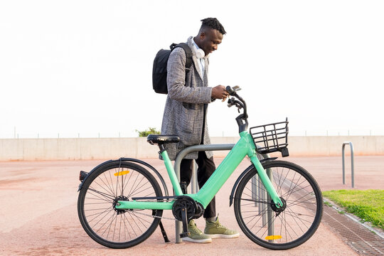Side view of African American male office employee using app on cellphone while parking bike in city