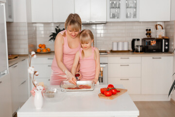 Mom and daughter cook in the kitchen. A young woman cooks with her child in the kitchen at home. Girls in pink clothes cook pizza. Happy and cheerful mom and daughter