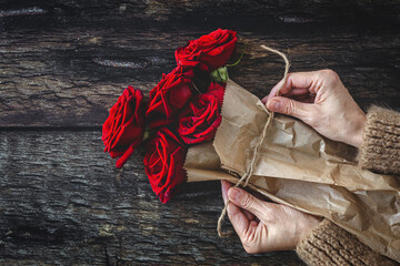 Top view of crop anonymous female in warm sweater wrapping bouquet of red roses with craft paper and rope against wooden background