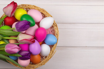 Basket with multi-colored Easter eggs and tulips on a white wooden background. View from above. Copy space. Festive spring banner