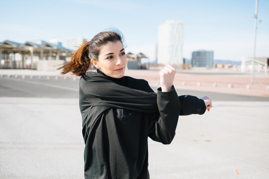Positive fit young female in black sportswear stretching arms while warming up on sunny road in suburb and looking away contentedly