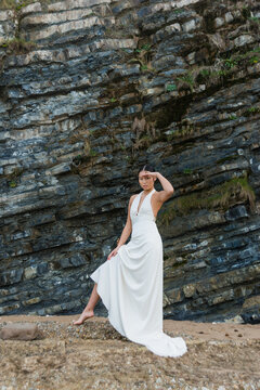 Full body of young slim barefoot Asian female in elegant white maxi dress keeping hand near forehead and looking at distance while standing near rough rocky formation