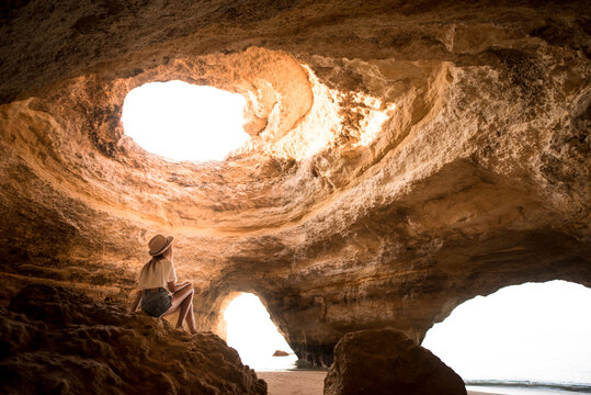 Side view of anonymous female in hat sitting in rock looking at hole in ceiling of cave near sea on sunny day in Algarve, Portugal