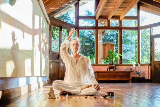 Young mindful male barefoot male sitting in lotus pose practicing yoga on floor near bowl gong and Buddha statuette