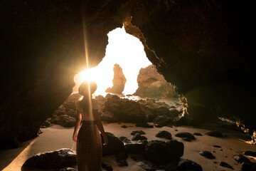 Back view of anonymous female in hat standing in rocky sand looking at entrance of cave near sea on sunny day in Algar seco caves in Algarve, Portugal