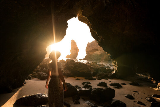 Back view of anonymous female in hat standing in rocky sand looking at entrance of cave near sea on sunny day in Algar seco caves in Algarve, Portugal