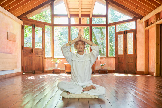 Young mindful male barefoot male sitting in lotus pose with eyes closed practicing yoga on floor near bowl gong and Buddha statuette