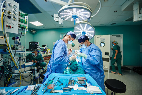Side view of unrecognizable young concentrated male assistant in sterile mask and gown giving instruments to plastic surgeon performing rhinoplasty in operating room