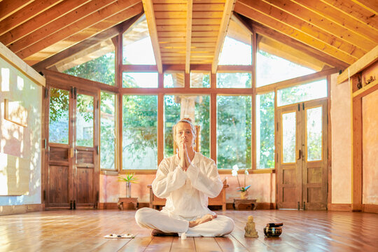 Young mindful male barefoot male sitting in lotus pose practicing yoga on floor near bowl gong and Buddha statuette
