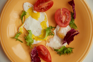 Morning meal with bright products: fried equal eggs, tomato and salad on bright orange plate. Top view