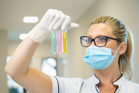 Serious female scientist in mask and latex gloves standing with glass test tubes filled with chemical liquids of various colors and working in lab