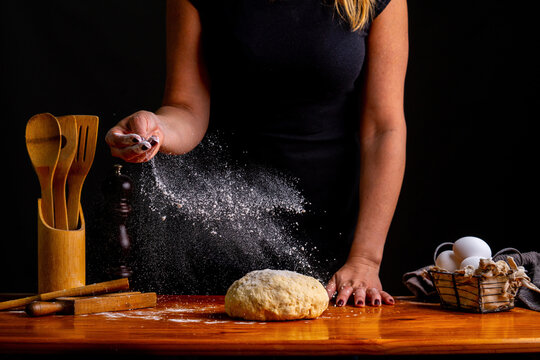 Crop female sprinkling kneaded bread dough with flour standing at wooden table with utensils and ingredients