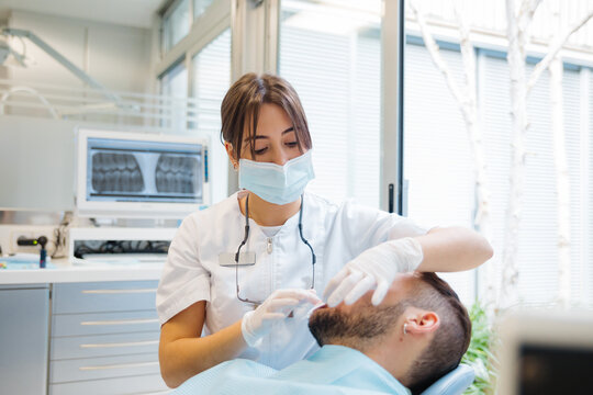 Concentrated professional female dentist in uniform and mask checking male patients teeth condition while working in modern dental clinic
