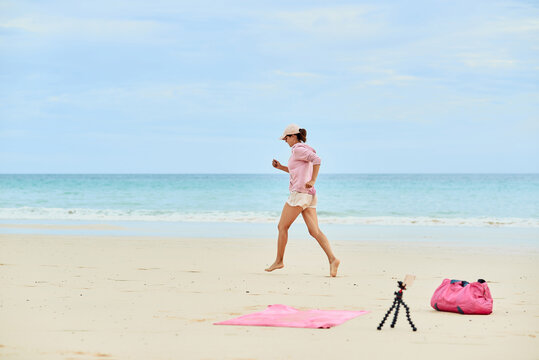 Side view full body of active female traveler running during training on sandy beach and recording video on mobile phone