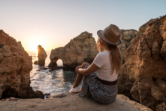 Side view of anonymous female in hat sitting on stone and admiring sea and cliffs at sundown in Ponta da Piedade in Algarve, Portugal