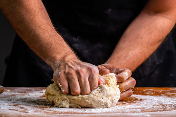 Crop man at wooden table kneading and rolling pile of dough for bread