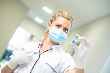 Female medic in protective mask and gloves standing with blue chemical liquid in glass vial in hospital during pandemic of coronavirus
