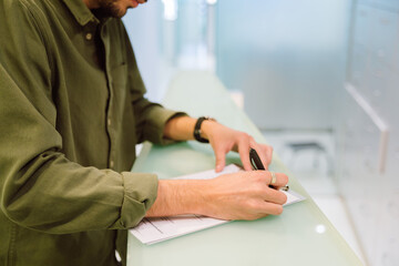 Side view crop focused male in casual clothes filling survey form while standing near clinic reception counter