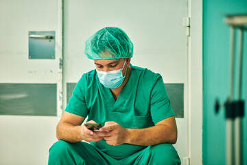 Anonymous young male doctor in green medical uniform and mask messaging on mobile phone while sitting in operating room after surgery
