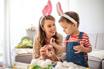 Cute boy sitting with his mother painting eggs for Easter. Mother and son enjoying while painting Easter eggs. Happy son and mother preparing creative eggs. Mother and son painting Easter eggs