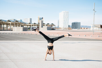 Back view full length fit determined sportswoman in tight activewear doing handstand exercise on asphalt road on sunny suburb with open legs