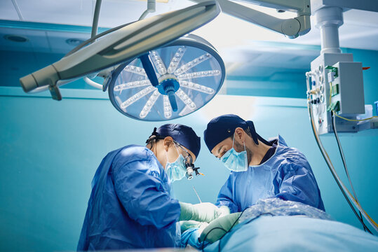Side view of unrecognizable male doctor with assistant in medical gowns and masks performing surgery with laser in operating room