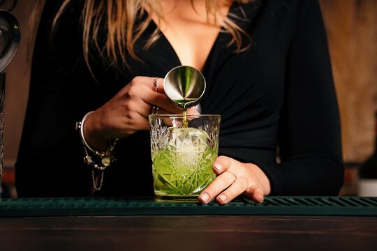 Crop unrecognizable female barkeeper poring green liquor into glass with ice cubes while preparing alcoholic cocktail in restaurant