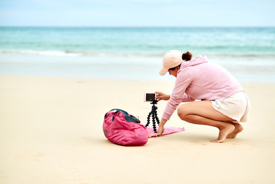 Side view full body of female travel blogger putting cellphone on tripod in sand for shooting video for social media