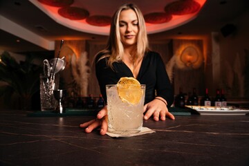 Content young female bartender with long blond hair serving cold alcohol cocktail decorated with lemon slice in stylish restaurant