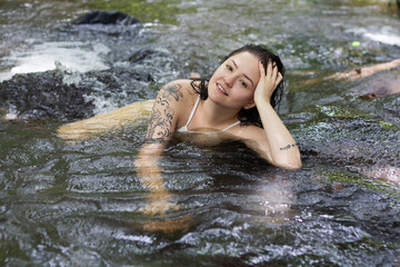 One white, tattooed and young woman smiling, relaxing and swimming outdoors on  with waterfall in nature, enjoying and having fun on her vacation