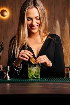 Self assured happy focused young female barkeeper with long blond hair in stylish outfit decorated cocktail with lemon slices while standing at counted in stylish bar