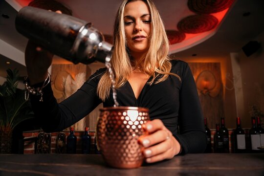 Beautiful blonde young female bartender in stylish outfit pouring alcohol cocktail from shaker into elegant metal mug in restaurant