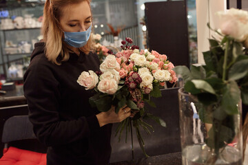 Florist works with colors. Flower seller chooses flowers for future bouquet. Flowers shop worker in a mask standing in flower shop and checking flowers in glass vase.