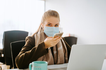 Blond business woman with mask working in front of computer in an office and using a cellphone