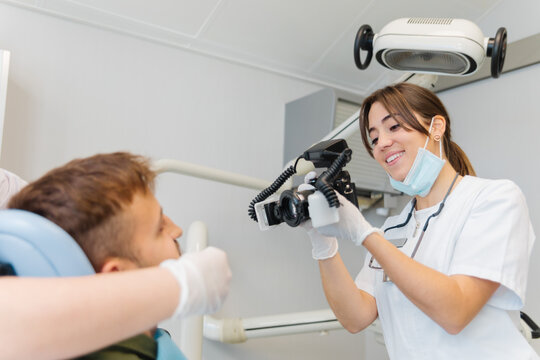 Smiling competent female dentist taking photos of male patient teeth while working in contemporary equipped dental clinic