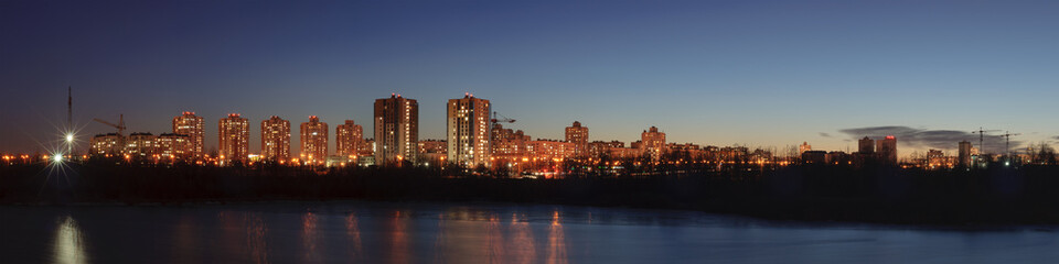 wide evening panoramic view of the modern residential quarter of the city in the coastal area with high-rise concrete buildings with light in the windows and bright street lighting
