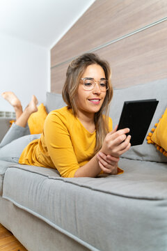 Smiling female lying on soft couch in living room and reading interesting e-book while spending time at home