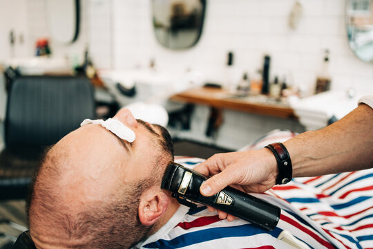 Crop anonymous master trimming beard of masculine man with electric machine in hairdressing salon