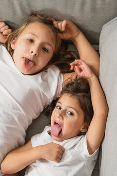 From above cheerful little sisters grimacing and showing tongues while lying on comfy couch and looking at camera
