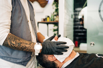 Side view of crop anonymous hipster male beauty master wrapping face of client in hot wet towel in barbershop