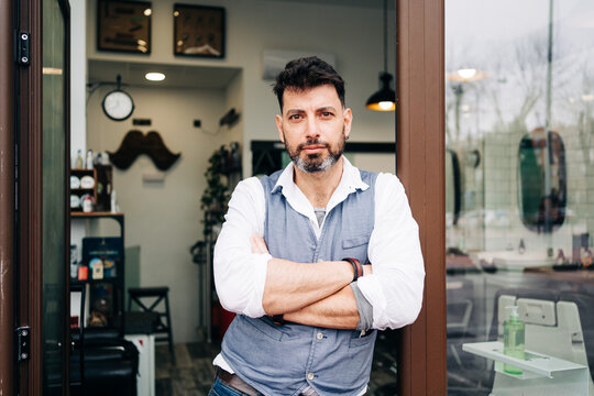 Adult bearded male beauty master with arms crossed looking at camera standing at door of barbershop