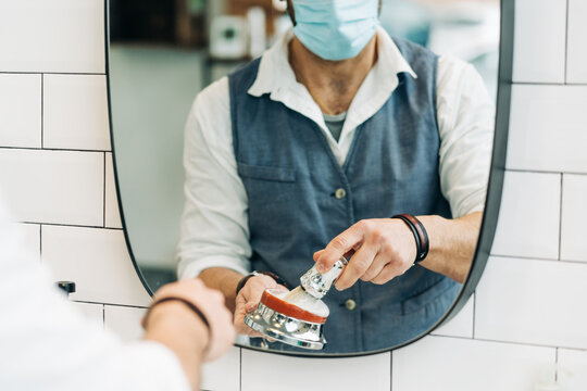 Crop anonymous male beauty master in sterile mask preparing shave brush with soap in bowl against mirror in bathroom at work