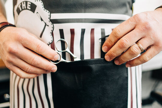 Crop unrecognizable male barber with scissors and comb in striped apron in hairdressing salon